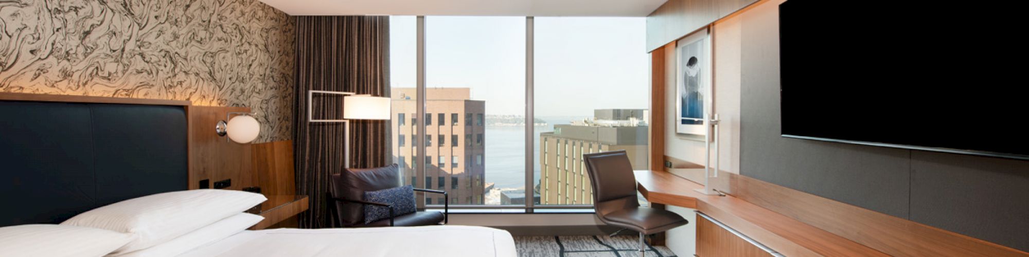 A modern hotel room with a large bed, a wall-mounted TV, a desk, a chair, and a window with a city view is shown.