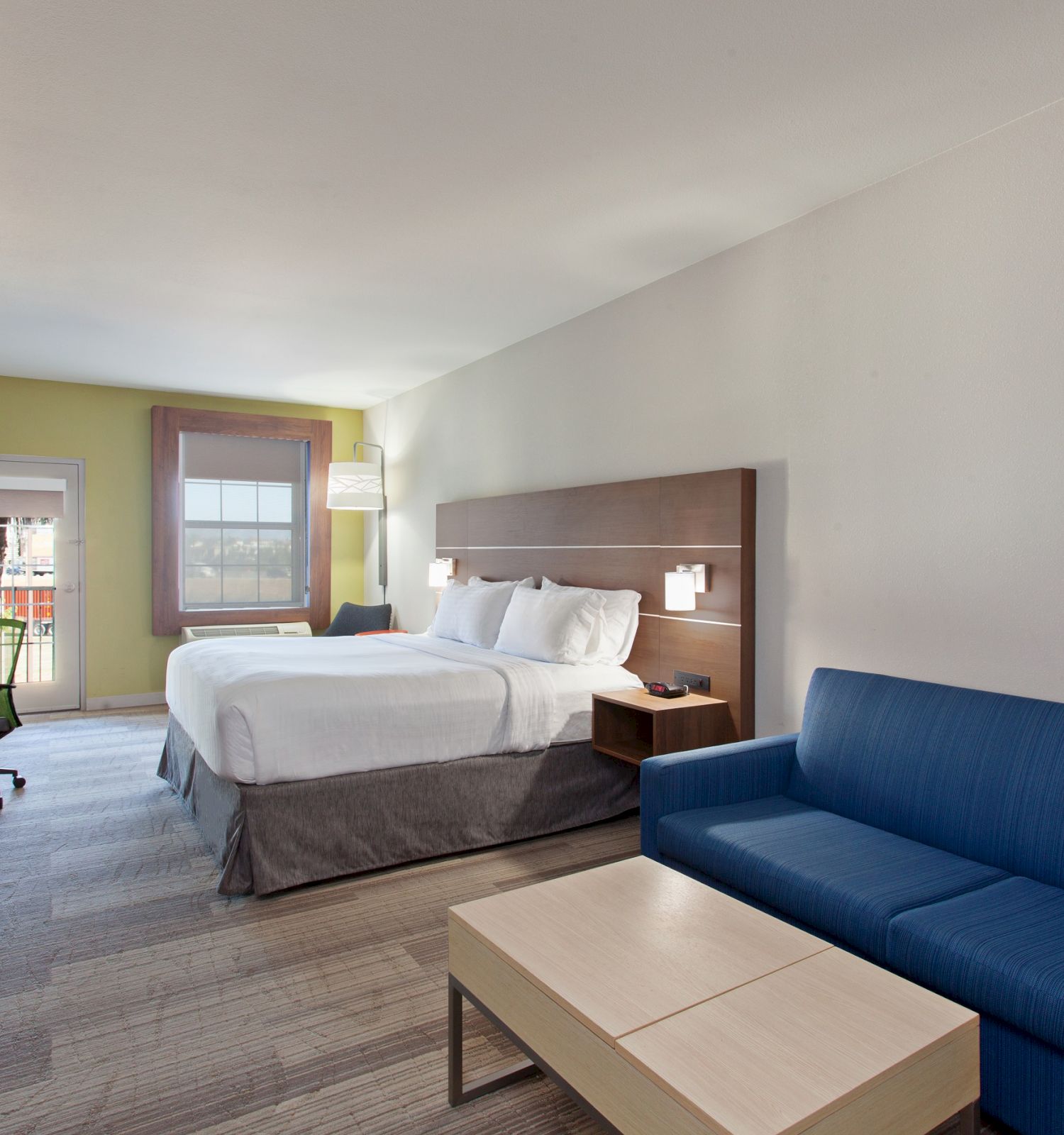 A modern hotel room with a king-size bed, blue sofa, coffee table, TV, desk with chair, and window providing natural light is shown.