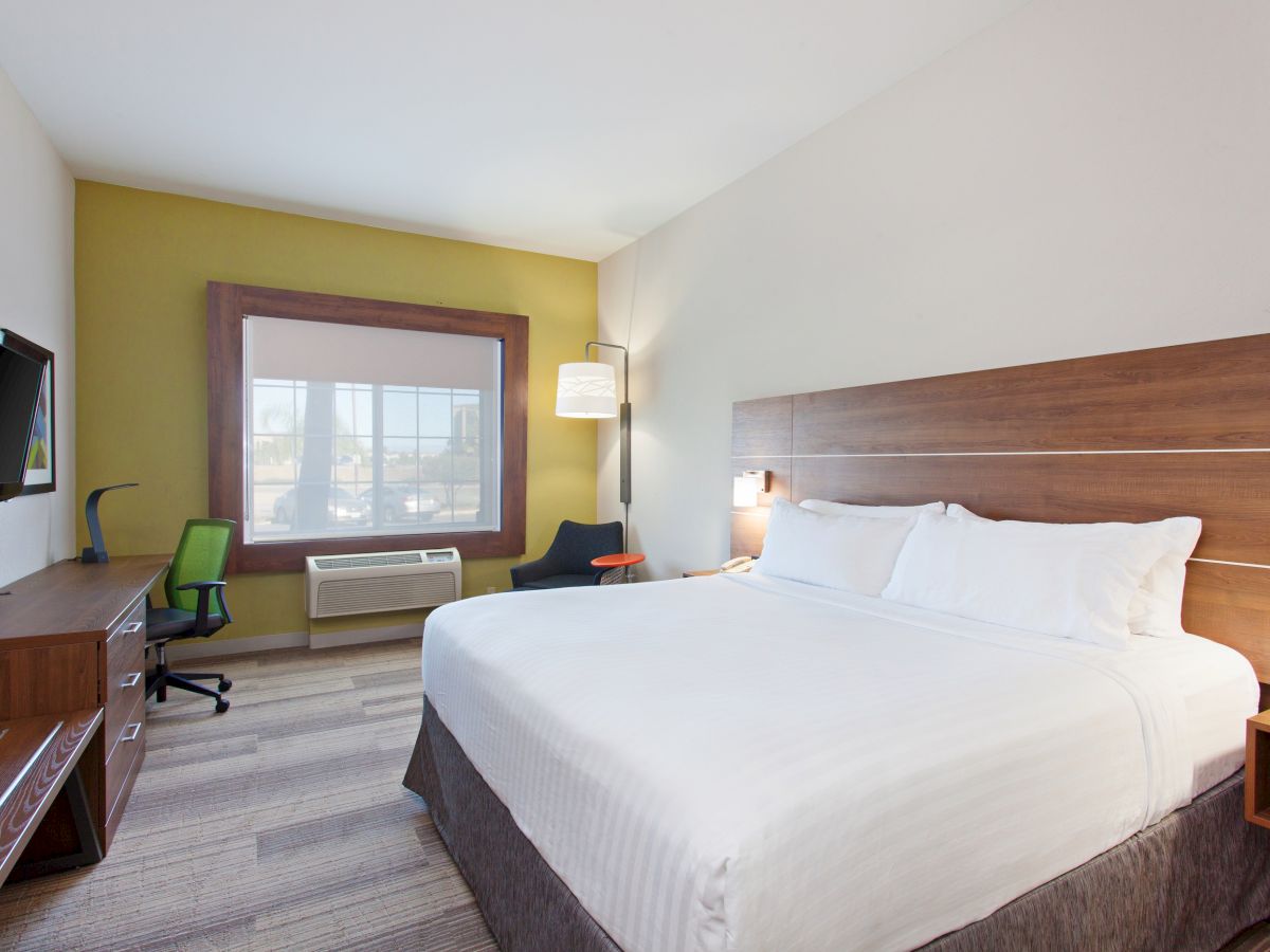 A hotel room with a king-sized bed, a desk with a TV, a desk chair, an armchair, and a window is shown.