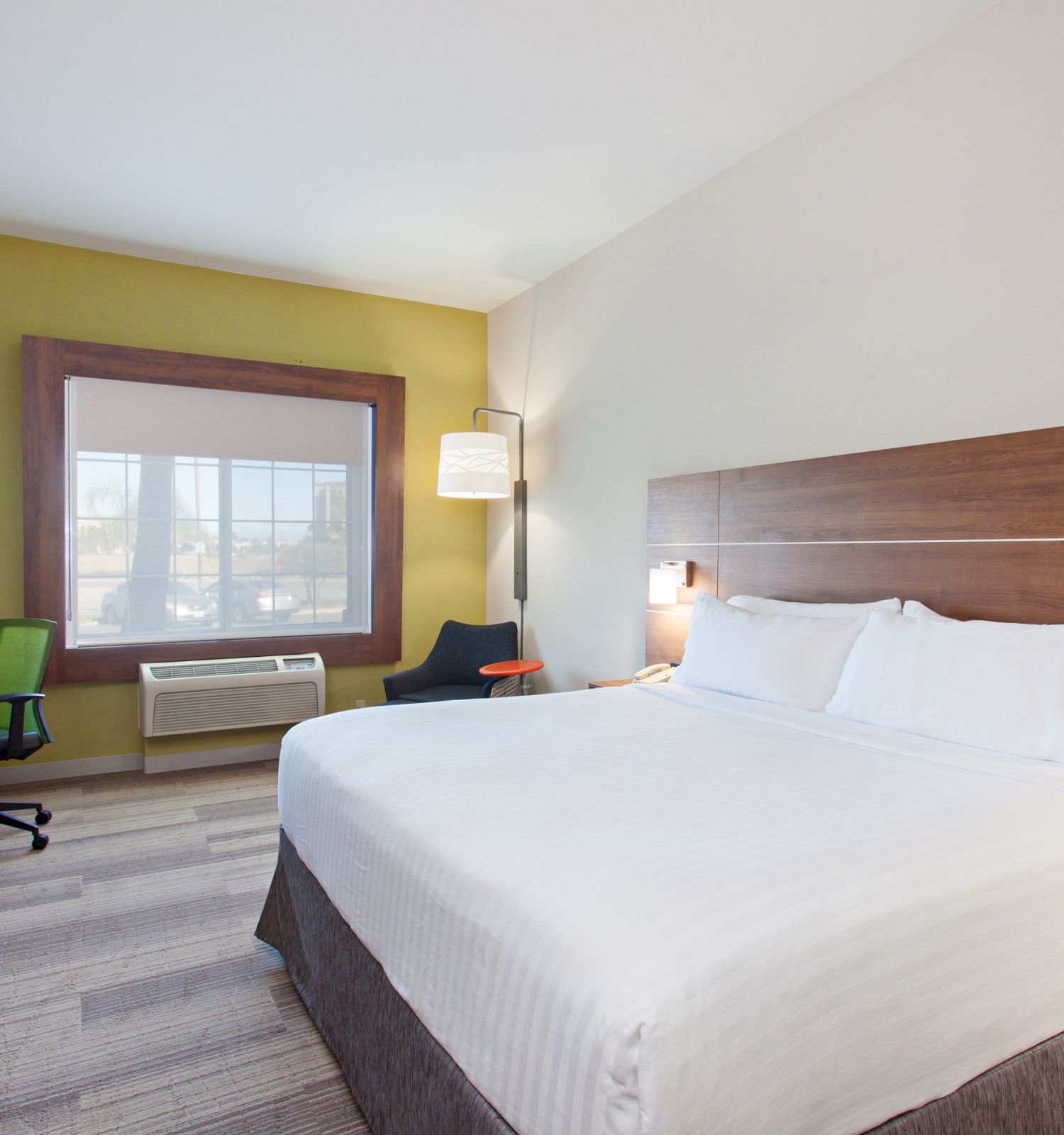 A hotel room with a king-sized bed, a desk with a TV, a desk chair, an armchair, and a window is shown.