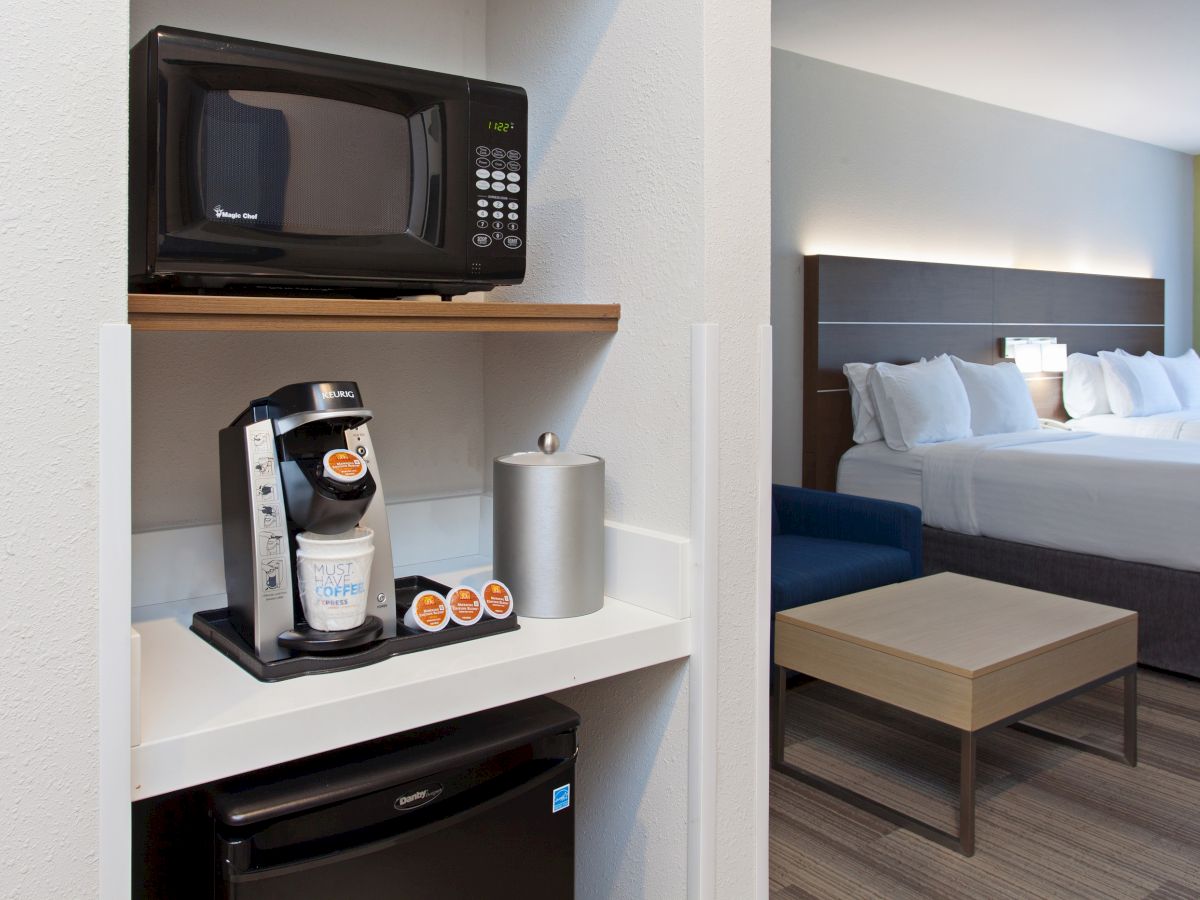 The image shows a hotel room with a microwave, coffee maker, and mini-fridge in the foreground and a bed in the background.