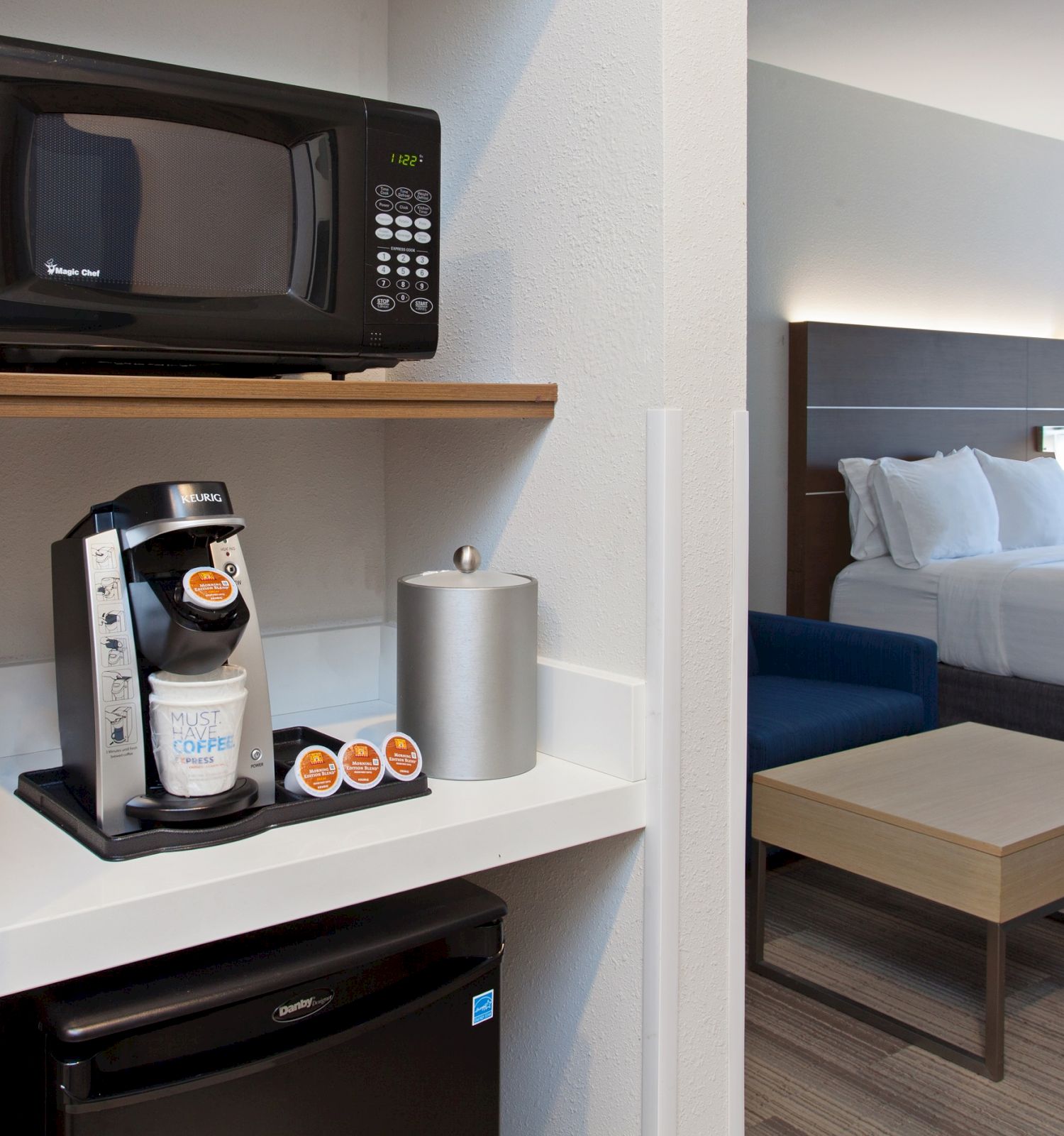 The image shows a hotel room with a microwave, coffee maker, and mini-fridge in the foreground and a bed in the background.