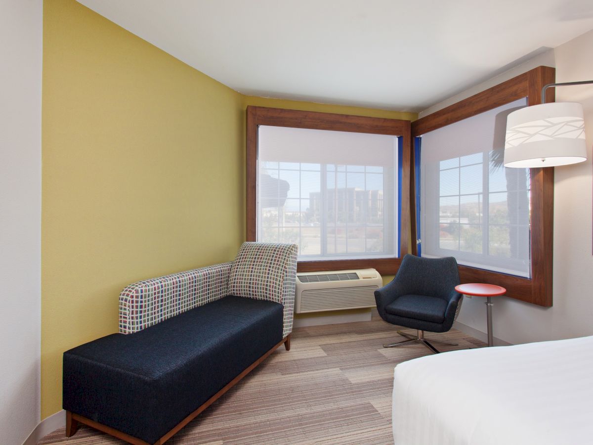 A modern room features a black chair, a small table, a sofa, and large windows with a view. The décor is minimal and contemporary.