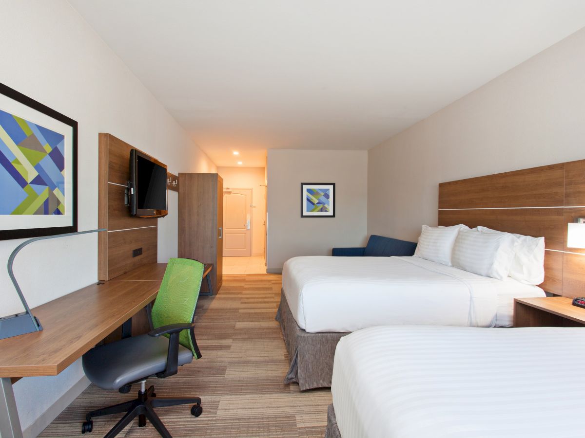 A hotel room with two beds, a wooden desk, green chair, wall art, TV, and overhead lighting, emitting a clean and modern vibe.