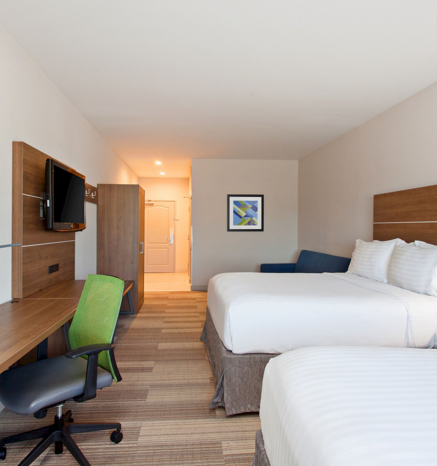 A hotel room with two beds, a wooden desk, green chair, wall art, TV, and overhead lighting, emitting a clean and modern vibe.