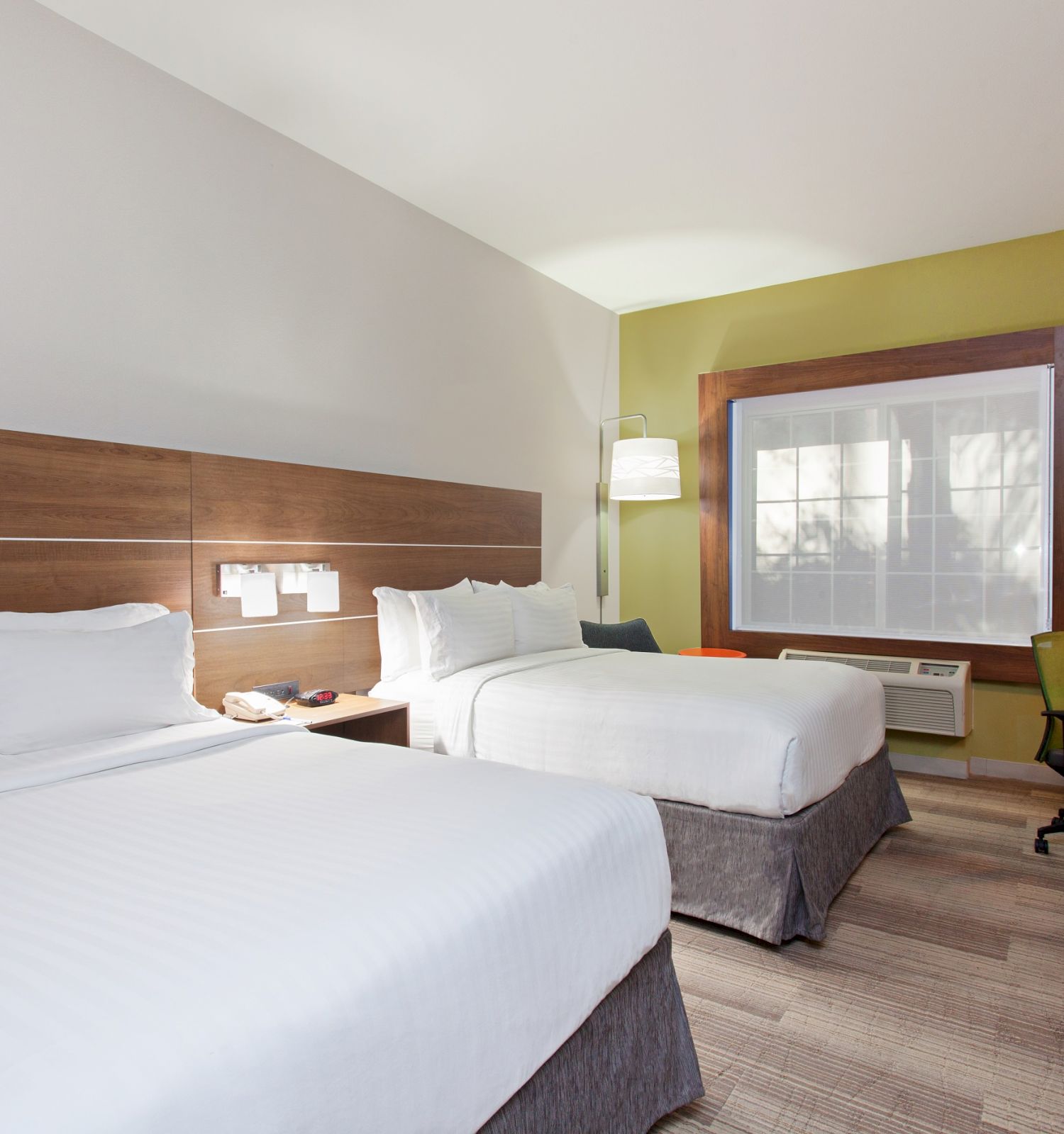 This image depicts a hotel room with two neatly made double beds, a desk with a lamp and chair, a TV, and a large window letting in natural light.