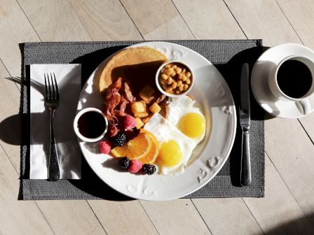 A breakfast plate with eggs, beans, toast, bacon, blackberries, oranges, and raspberries. A fork, knife, napkin, and cup of coffee are also visible.