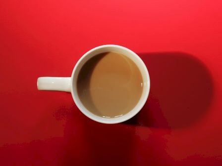 A white coffee cup filled with coffee is placed on a red surface, casting a shadow to its right in the image.